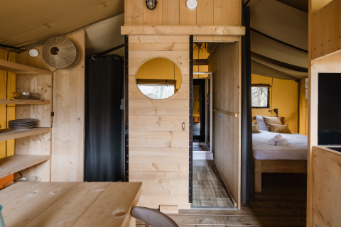 Vue intérieure d'une tente safari avec meubles en bois, étagères ouvertes, table à manger et chambre.