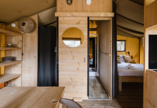 Vue intérieure d'une tente safari avec meubles en bois, étagères ouvertes, table à manger et chambre.