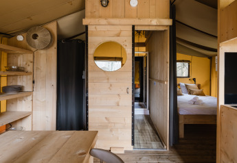 Innenraum eines Safarizeltes mit Holzmöbeln, Schlafzimmer, offenen Regalen und gemütlicher Atmosphäre.