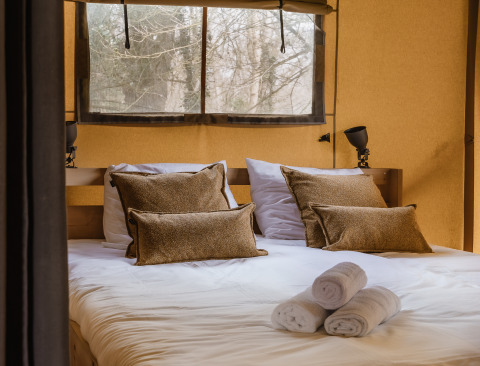 Lit double confortable avec coussins marron et serviettes blanches dans une tente safari, vue sur la forêt.