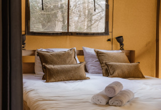 Lit double confortable avec coussins marron et serviettes blanches dans une tente safari, vue sur la forêt.