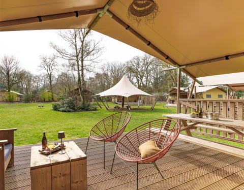 Vue depuis la terrasse d'une tente safari avec chaises confortables, table et pelouse verte en pleine nature.