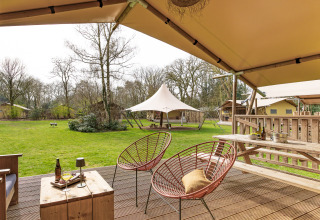 Vue depuis la terrasse d'une tente safari avec chaises confortables, table et pelouse verte en pleine nature.