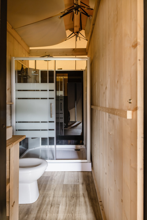 Salle de bain dans une tente safari avec murs en bois, douche en verre, toilettes et sol stratifié.