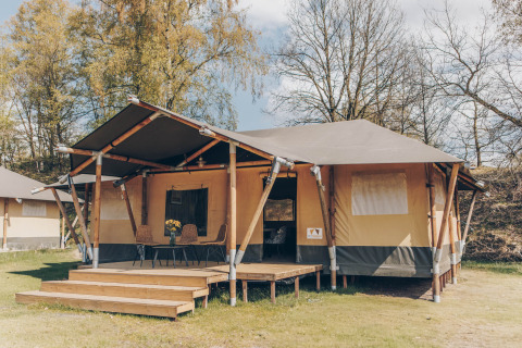 Een safaritent met houten veranda en stoelen ervoor, omgeven door groene bomen en natuur.