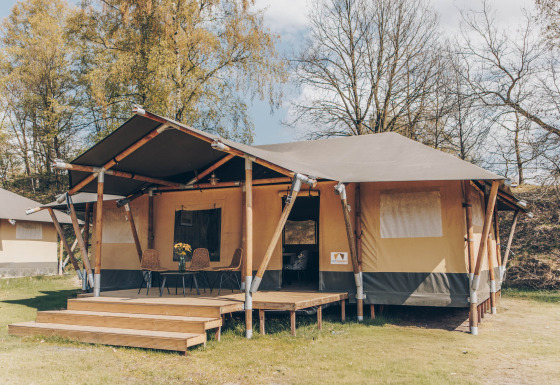 Een safaritent met houten veranda en stoelen ervoor, omgeven door groene bomen en natuur.