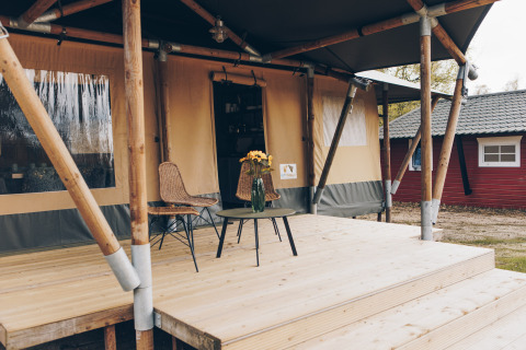 Buitenveranda van een safaritent met twee stoelen, een tafeltje en een vaas bloemen op hout.