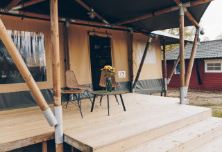 Buitenterras van een safaritent met twee stoelen, een tafel en een vaas bloemen op houten vloer.