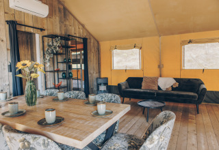 Interieur van een safaritent met houten wanden, eettafel, comfortabele bank en bloemen in een vaas.