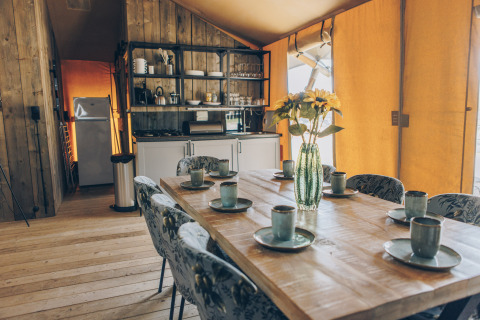 Safaritent interieur met houten eettafel voor zes, bloemenvaas, stoelen en kleine, open keukenruimte.