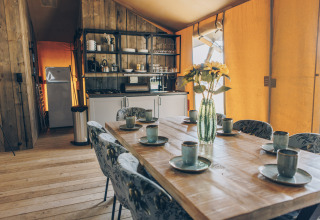 Interieur van safaritent met houten eettafel gedekt voor zes, bloemenvaas en knusse open keukenopstelling.