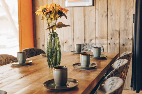 Houten tafel met keramieken mokken, borden en een groene vaas met bloemen in een safaritent met houten wanden.