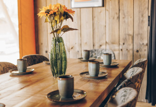Houten tafel met keramieken mokken, borden en een groene vaas met bloemen in een safaritent met houten wanden.