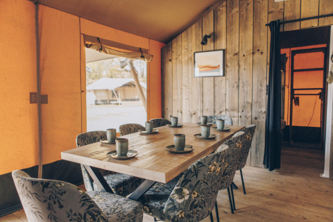 Interieur van een safaritent met houten tafel, zes stoelen met bloemenprint en keramische bekers op tafel.