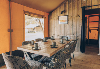Binnenzicht van een safaritent met houten tafel, zes bloemstoelen en keramieken bekers klaar voor gebruik.