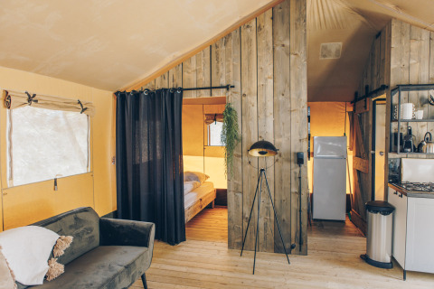 Interieur van een safaritent met gezellige zithoek, keuken, slaapkamer en rustieke houten wanden.