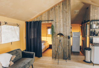 Interieur van een safaritent met gezellige zithoek, keuken, slaapkamer en rustieke houten wanden.