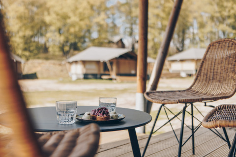 Twee glazen water en een dessert op een tafel bij rotan stoelen, met safaritenten op de achtergrond.