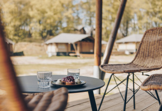 Twee glazen water en een toetje op een tafel naast rotan stoelen, met safaritenten op de achtergrond.