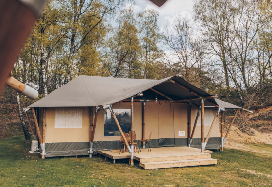 Safaritent Glamfield Lodge met houten veranda en stoelen, omgeven door bomen en groene natuur.