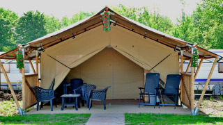 Safari tent with toilet, featuring comfy chairs and flowers on the porch, set in a lush campground.