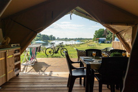 View from inside a safari tent with wooden deck, outdoor furniture, bicycle, lake, and picnic tables.