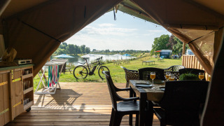 Vista dall'interno di una tenda safari con terrazza in legno, bicicletta, lago e tavoli da picnic.