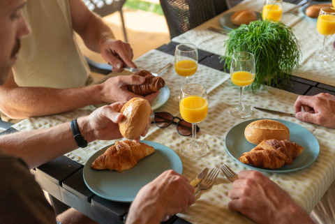 Quattro persone fanno colazione con croissant, panini e succo d'arancia in una tenda safari al campeggio.