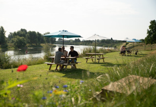 People sitting at picnic tables with umbrellas by the river at Campsite de Boomgaard, Safari Tent Premium Maaszicht.