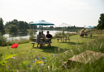 Menschen sitzen an Picknicktischen mit Sonnenschirmen am Flussufer des Campingplatzes de Boomgaard in Belgien.