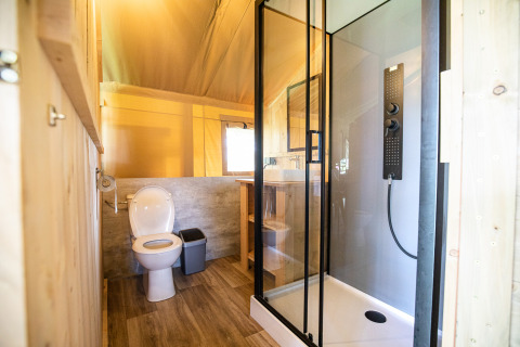 Moderne badkamer in een safaritent met douche, toilet, raam en houten accent onder natuurlijk licht.