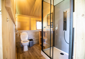 Modern bathroom inside a safari tent featuring a shower, toilet, window, and wood accents in daylight.