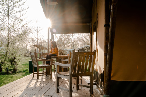 Person sitzt auf einer Terrasse vor einem Safarizelt mit Holzmöbeln auf Campingplatz De Rammelbeek, Niederlande.