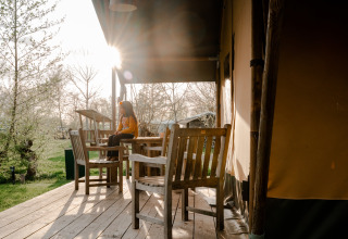 Person sitzt auf einer Terrasse vor einem Safarizelt mit Holzmöbeln auf Campingplatz De Rammelbeek, Niederlande.