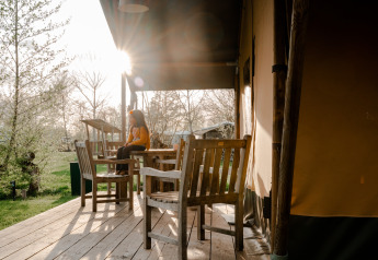 Person sitzt auf einer Terrasse vor einem Safarizelt mit Holzmöbeln auf Campingplatz De Rammelbeek, Niederlande.