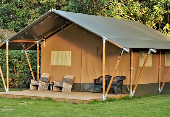 Safari-Zelt mit hölzerner Terrasse, Stühlen davor und üppigem Grün sowie Bäumen im Hintergrund.