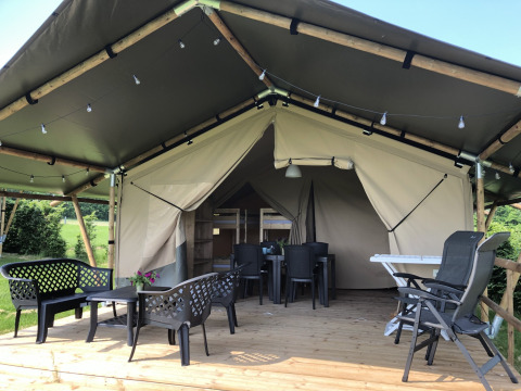 Tenda safari con veranda in legno, sedie all’aperto, luci e circondata da verde paesaggio.