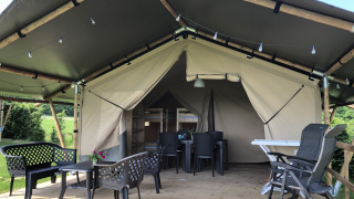 Tenda safari con veranda in legno, sedie all’aperto, luci e circondata da verde paesaggio.