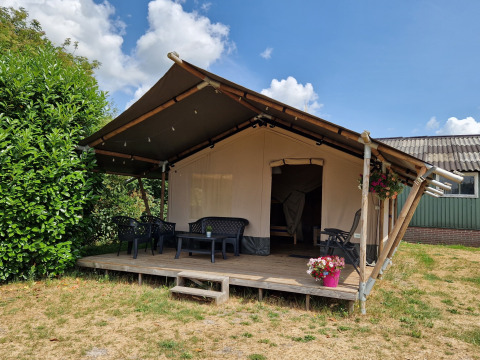 Safari telt med træveranda, plastikstole og blomsterpotte under en blå himmel med skyer.