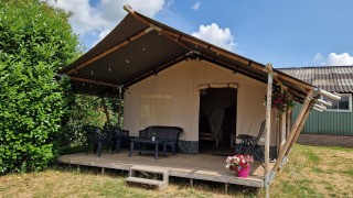 Safari telt med træveranda, plastikstole og blomsterpotte under en blå himmel med skyer.
