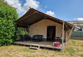 Safari tent with a wooden veranda, plastic chairs, and a flower pot under a blue sky with clouds.