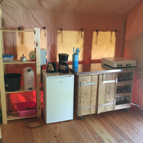 Keukenhoek in een safaritent met koelkast, koffiezetapparaat, fornuis en open houten kast op een houten vloer.