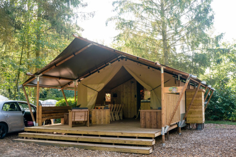 Tenda safari con servizi igienici, terrazza in legno, posti a sedere e ambiente naturale boscoso.