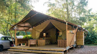 Tenda safari con servizi igienici, terrazza in legno, posti a sedere e ambiente naturale boscoso.