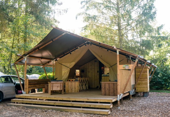 Tenda safari con servizi igienici, terrazza in legno, posti a sedere e ambiente naturale boscoso.