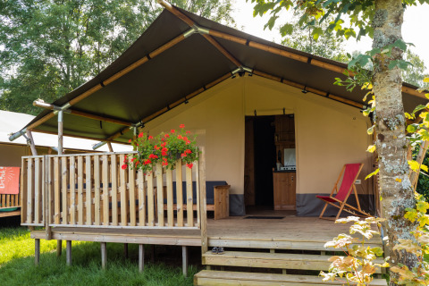 Tenda safari con servizi igienici, terrazza in legno, fiori e sedia sdraio circondata dal verde.