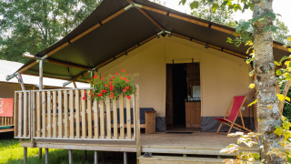 Tenda safari con servizi igienici, terrazza in legno, fiori e sedia sdraio circondata dal verde.
