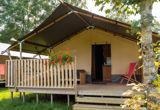 Tenda safari con servizi igienici, terrazza in legno, fiori e sedia sdraio circondata dal verde.