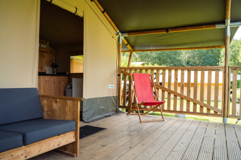 Veranda di tenda safari con divano in legno, sedia rossa e vista sul verde paesaggio naturale.