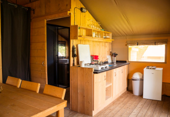 Interieur van een safaritent met houten eettafel, compacte keuken, koelkast en een raam.